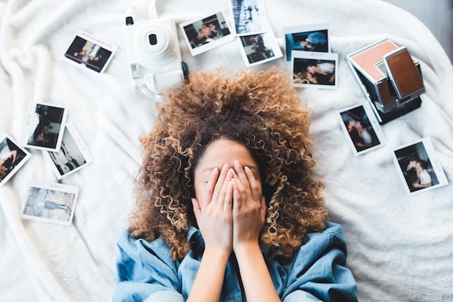 A woman lies on her bed surrounded by photos, while covering her face